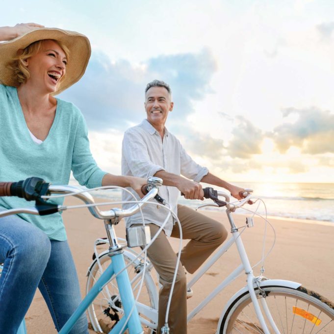 Mature couple cycling on the beach at sunset or sunrise. Mature couple cycling on the beach at sunset or sunrise. They are laughing and having fun. They are casually dressed. Could be a retirement vacation.